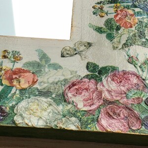 May include: A decorative mirror with a floral design. The frame is painted white and green, with embossed roses, pansies, and butterflies. The flowers are in shades of pink, red, and purple, with green leaves. The mirror is a vintage style.