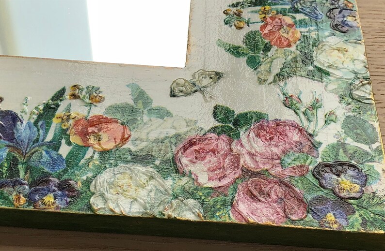 May include: A decorative mirror with a floral design. The frame is painted white and green, with embossed roses, pansies, and butterflies. The flowers are in shades of pink, red, and purple, with green leaves. The mirror is a vintage style.