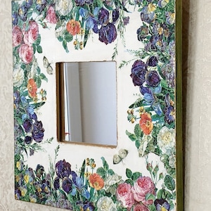 May include: Square decorative mirror with a floral design featuring roses, irises, and butterflies. The frame is white with a gold trim and green edges. The mirror is square with a clear reflection.
