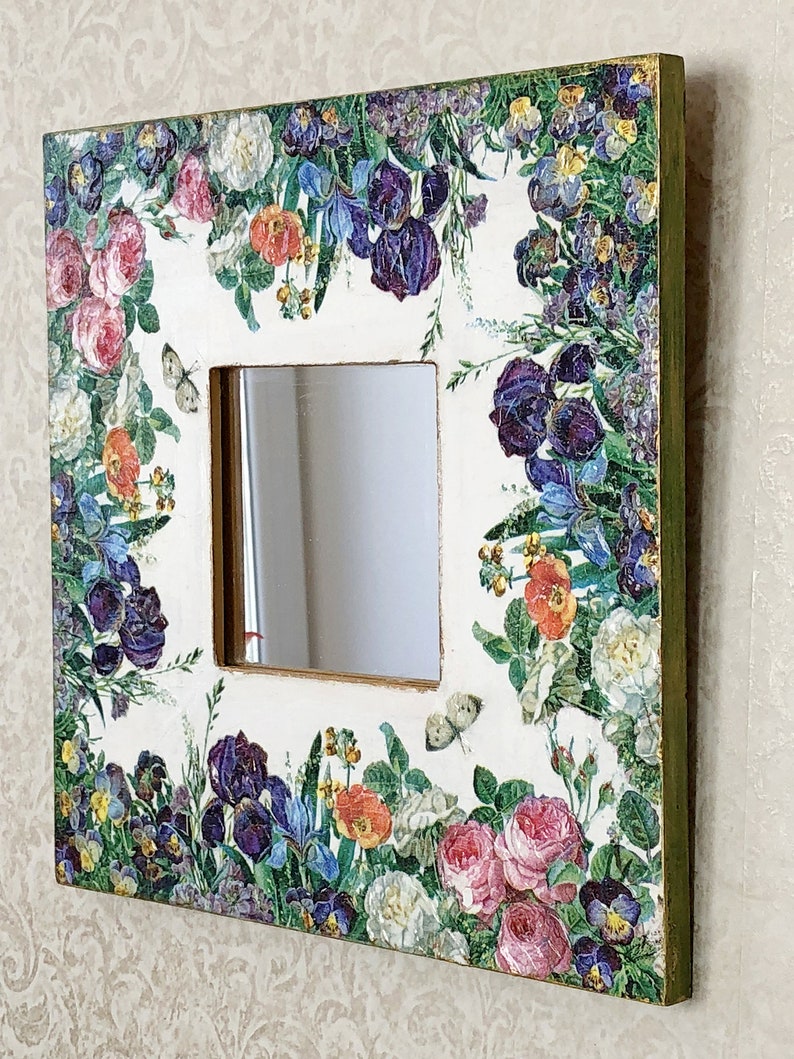 May include: Square decorative mirror with a floral design featuring roses, irises, and butterflies. The frame is white with a gold trim and green edges. The mirror is square with a clear reflection.