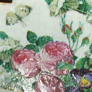 May include: Close-up of a decorative floral design featuring roses, buds, and a butterfly. The image showcases a mix of colors, including pink, white, green, and purple, with a glossy finish. The design is reminiscent of vintage botanical art.