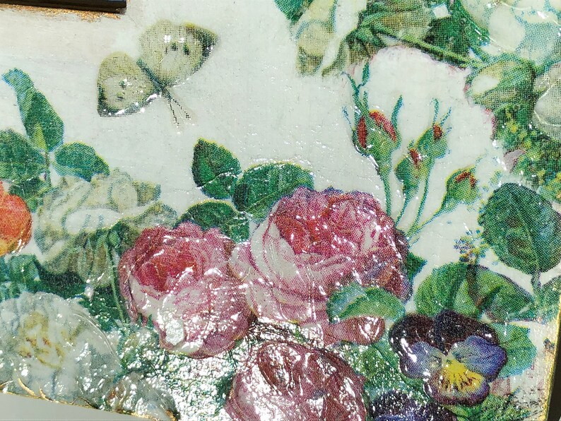 May include: Close-up of a decorative floral design featuring roses, buds, and a butterfly. The image showcases a mix of colors, including pink, white, green, and purple, with a glossy finish. The design is reminiscent of vintage botanical art.
