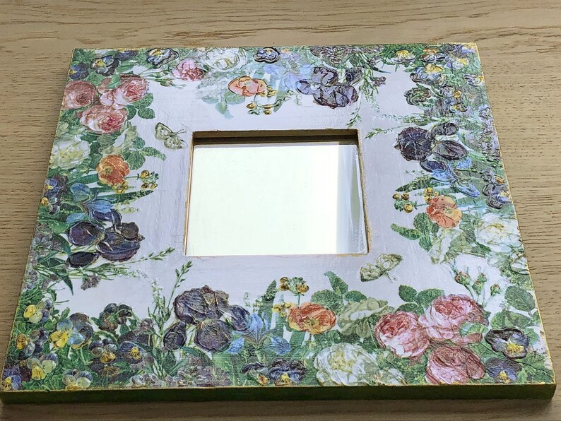 May include: Square decorative mirror with a floral design. The frame is white with a green edge and is covered in a pattern of colorful flowers, including roses, irises, and pansies. Butterflies are also featured in the design.