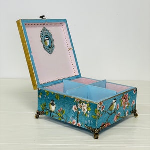 Puede incluir: Una caja de madera cuadrada con la tapa abierta, que presenta un exterior azul con un patr&oacute;n de flores y p&aacute;jaros. El interior est&aacute; dividido en cuatro secciones con paredes azul claro y una base rosa p&aacute;lido. Se apoya sobre cuatro patas doradas.