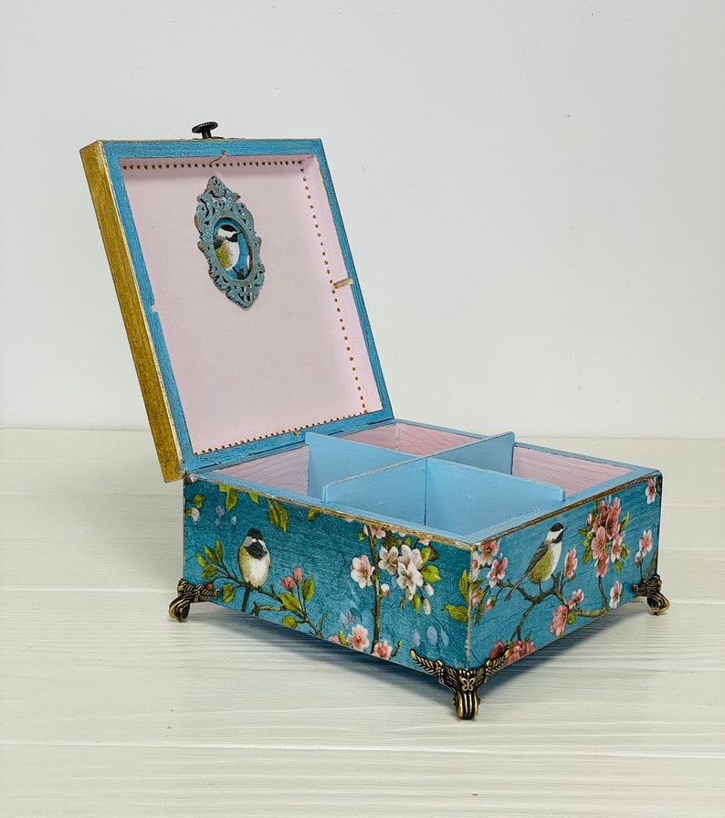 Puede incluir: Una caja de madera cuadrada con la tapa abierta, que presenta un exterior azul con un patr&oacute;n de flores y p&aacute;jaros. El interior est&aacute; dividido en cuatro secciones con paredes azul claro y una base rosa p&aacute;lido. Se apoya sobre cuatro patas doradas.