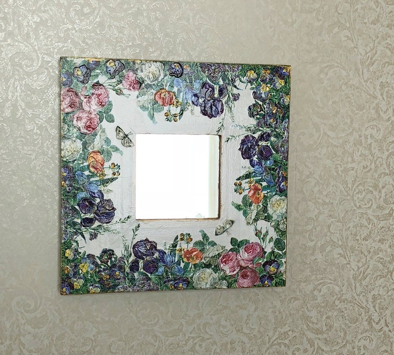 May include: Square decorative mirror with a floral design. The frame is white with a collage of colorful flowers, including roses and irises, and butterflies. The mirror is square and has a gold trim.