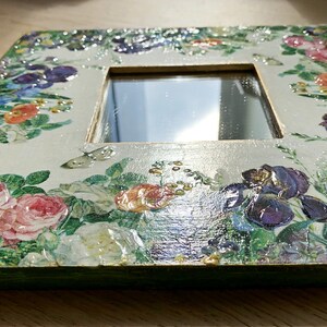 May include: Square decorative mirror with a floral design. The frame is decorated with a variety of flowers in shades of pink, purple, and white, with green leaves. The mirror is surrounded by a cream-colored border.