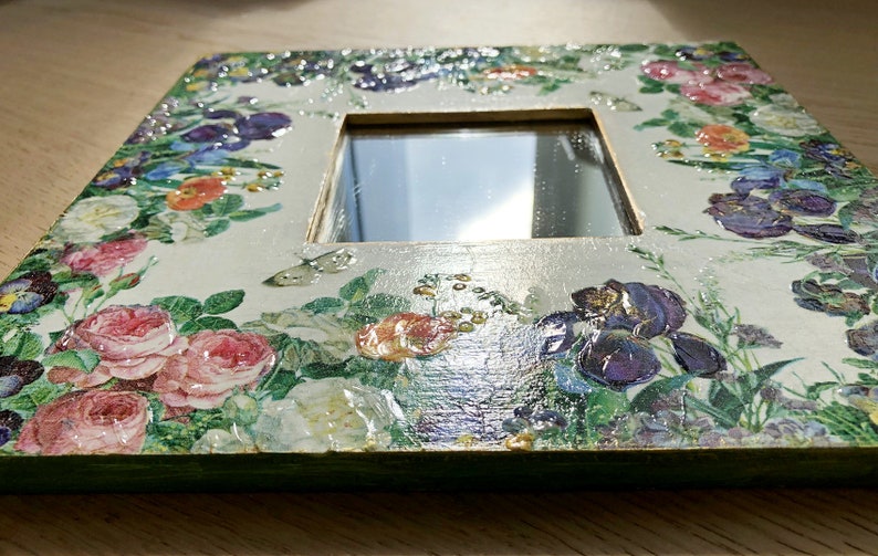 May include: Square decorative mirror with a floral design. The frame is decorated with a variety of flowers in shades of pink, purple, and white, with green leaves. The mirror is surrounded by a cream-colored border.