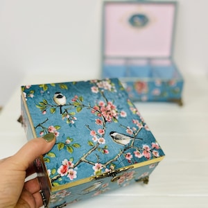 Puede incluir: Una caja decorativa cuadrada con fondo azul y un patr&oacute;n de flores de cerezo rosas y p&aacute;jaros peque&ntilde;os. La caja tiene patas y un cierre de metal dorado. Una segunda caja abierta con interior rosa claro es visible en el fondo.