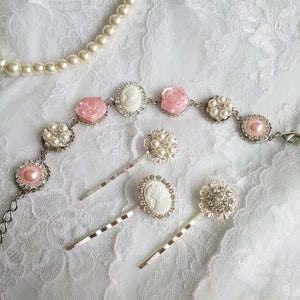 May include: A collection of vintage-style jewelry on a white lace surface. The items include a pearl necklace, a bracelet with pink rose and cameo accents, and several hair pins with pearl and crystal embellishments.