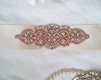 May include: A rose gold bridal sash with a beaded design featuring clear and pink rhinestones. The sash is on a white lace background.