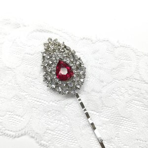 May include: A silver hair clip with a large red rhinestone in the center surrounded by smaller clear rhinestones. The clip is on a white lace background.