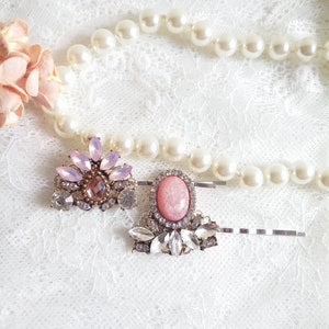 May include: Two hair clips with pink and clear rhinestones. The clips have a silver metal base and feature a large pink oval-shaped stone in the center.