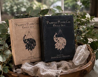Może przedstawiać: Dwa prostokątne pudełka na karty wyroczni zatytułowane Palimpsest Playing Card Oracle Deck stworzone przez Jasona i Lisę Williamsów. Jedno pudełko jest jasnobrązowe z czarną ilustracją pawia, a drugie czarne z jasnobrązową ilustracją pawia.