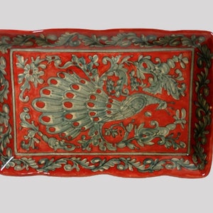 Può includere: Un vassoio rettangolare in ceramica rossa con un disegno di pavone. Il vassoio presenta un bordo smerlato e manici. Il pavone è raffigurato in tonalità di grigio e verde, circondato da motivi floreali e fogliari.