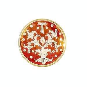 Peut inclure: Une assiette décorative ronde avec un bord doré. L'assiette présente un fond orange rougeâtre avec un motif floral blanc au centre et la lettre "T" en haut. De petits points blancs sont dispersés sur le bord de l'assiette.