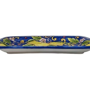 May include: A rectangular ceramic serving dish with a white base and a vibrant blue rim. The dish is decorated with a floral pattern featuring yellow, green, and white flowers and leaves. The design is symmetrical, with the floral elements arranged along the edges.