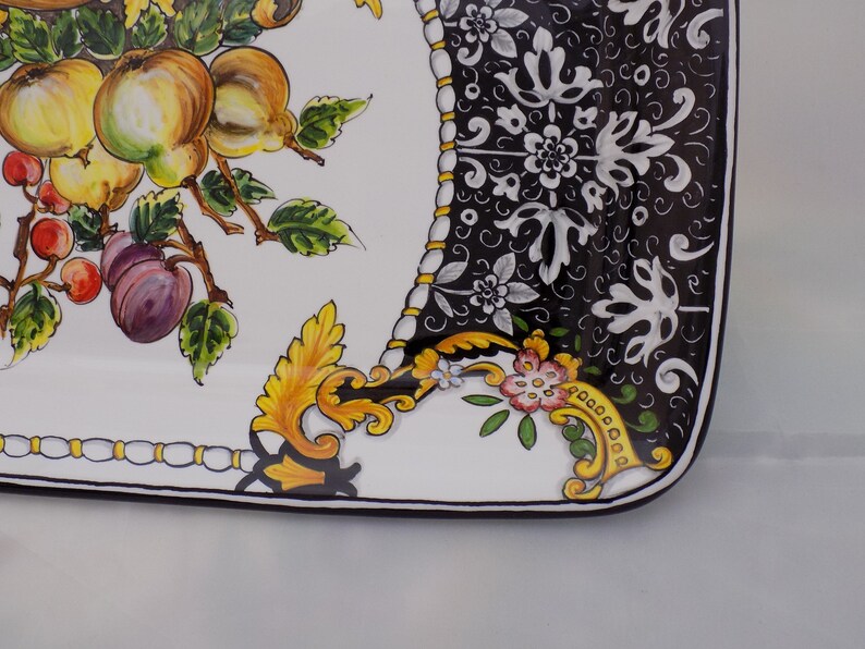 Puede incluir: Plato rectangular de cer&aacute;mica con fondo blanco y dise&ntilde;o pintado de frutas y flores. El dise&ntilde;o incluye manzanas amarillas, cerezas rojas y ciruelas moradas. El borde presenta un patr&oacute;n floral en blanco y negro con detalles dorados.