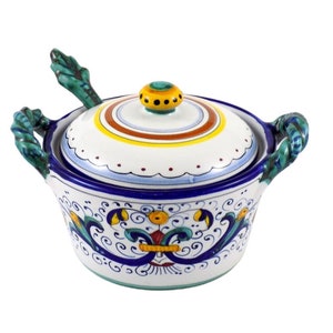 May include: A white ceramic soup tureen with a lid and spoon. The tureen has blue, yellow, and green decorative patterns. The handles and spoon are green. The lid has a yellow and green knob.