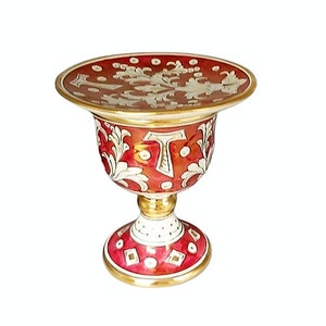 Może przedstawiać: Ozdobna, bogato zdobiona misa na cokole z szerokim, okrągłym blatem i rozkloszowanym, pucharowatym korpusem. Misa jest głównie czerwona ze złotymi wykończeniami i białymi wzorami kwiatowymi i geometrycznymi.