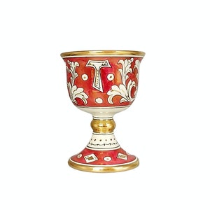 Op de afbeelding: Een decoratieve keramische kelk met een rood en wit bloemmotief. De kelk heeft een gouden rand en voet, met de letter "T" prominent op de voorkant. De kelk is ongeveer 13 cm hoog.