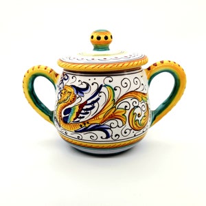May include: A white ceramic sugar bowl with a lid, featuring a colourful hand-painted design of a mythical creature with yellow, blue, green, and red accents. The bowl has two handles and a round shape.