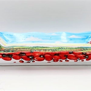 May include: Rectangular ceramic serving dish with a painted landscape scene and a border of red poppies. The interior features a blue sky, green fields, and a village. The dish is white on the bottom and has a wavy rim.