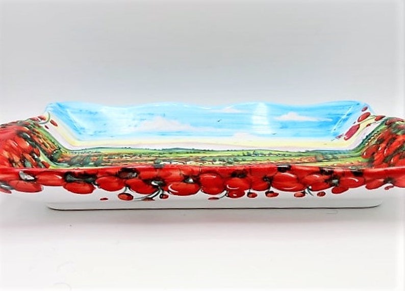 May include: Rectangular ceramic serving dish with a painted landscape scene and a border of red poppies. The interior features a blue sky, green fields, and a village. The dish is white on the bottom and has a wavy rim.