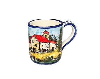 Puede incluir: Una taza de cerámica con una escena de paisaje pintada a mano. La taza presenta un edificio blanco con un tejado rojo, un cielo azul y árboles verdes. El asa es blanca con detalles azules y el borde es azul.