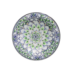 Puede incluir: Un plato redondo de cerámica con un diseño floral detallado. El plato presenta un patrón central en forma de flor en tonos verdes, azules y blancos, rodeado por un borde de intrincados motivos de hojas y vides. El borde es negro.