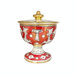 Op de afbeelding: Een decoratieve keramische kelk met deksel, rode achtergrond en witte bloemen en geometrische patronen. De kelk heeft een goudkleurige rand, voet en knop. De kelk wordt waarschijnlijk gebruikt voor decoratieve doeleinden.