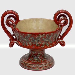 Puede incluir: Un cuenco de pedestal de cerámica roja con asas ornamentadas y un patrón floral decorativo. El cuenco tiene un interior beige y una base detallada. Las asas están enrolladas y añaden un elemento de diseño único.
