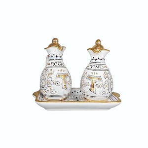 Puede incluir: Juego de aceitera y vinagrera de cerámica blanca con detalles dorados, que incluye dos botellas decorativas y una bandeja a juego. Las botellas están etiquetadas con "ACQUA" y "VINO" en escritura negra, con adornos dorados.