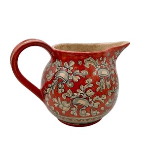 Könnte beinhalten: Eine rote Keramikkanne mit einem floralen Muster in Grau. Die Kanne hat einen runden Körper und einen gebogenen Griff.