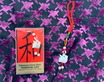 Może przedstawiać: Czerwone pudełko z chińskimi znakami i tekstem "Chinese Feng Shui PEACEFUL LIFE Hand Carved Jade Luck Charm" leży na wzorzystym tle. Czerwony sznurek z białym urokiem i koralikami wisi w pobliżu.