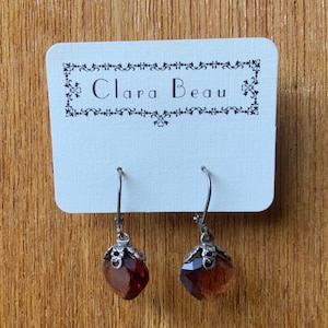 May include: A pair of silver dangle earrings with a dark red faceted gemstone. The earrings are on a white card with the text "Clara Beau" in black.