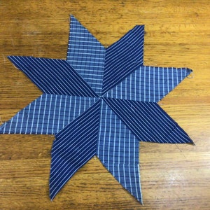 May include: A blue and white striped fabric star with eight points. The star is made of two different fabrics, one with a white and blue plaid pattern and the other with a white and blue striped pattern.
