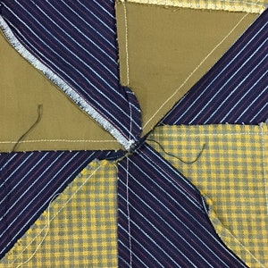 May include: Close-up of a quilt block with a star pattern. The block is made of four different fabrics: a yellow and green plaid, a blue and white striped fabric, and two pieces of a solid brown fabric. The fabrics are sewn together to create a star shape.