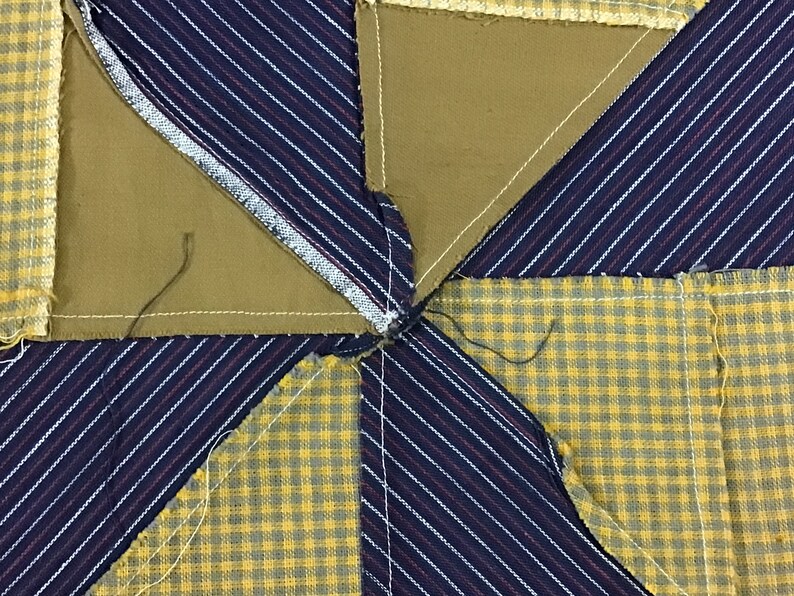 May include: Close-up of a quilt block with a star pattern. The block is made of four different fabrics: a yellow and green plaid, a blue and white striped fabric, and two pieces of a solid brown fabric. The fabrics are sewn together to create a star shape.