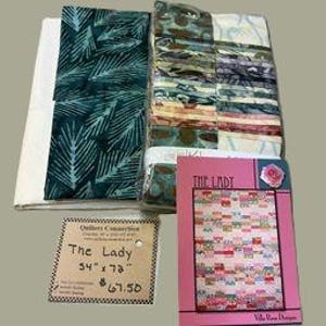 May include: A quilting fabric kit titled The Lady, featuring a stack of assorted fabric squares in shades of green, brown, and cream, a large piece of dark green leaf-patterned fabric, and a pattern booklet with a pink cover and quilt image.