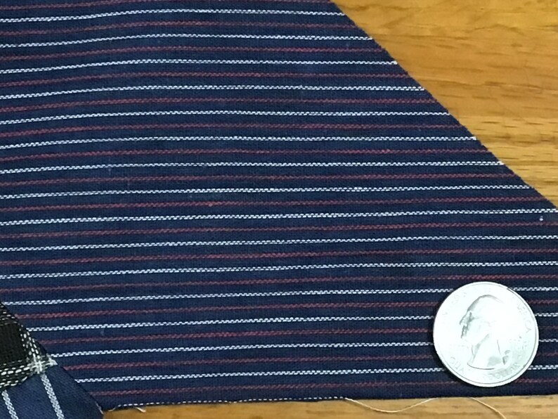 May include: Close-up of a dark blue fabric with thin white and red stripes. A silver coin is visible in the bottom right corner of the image.