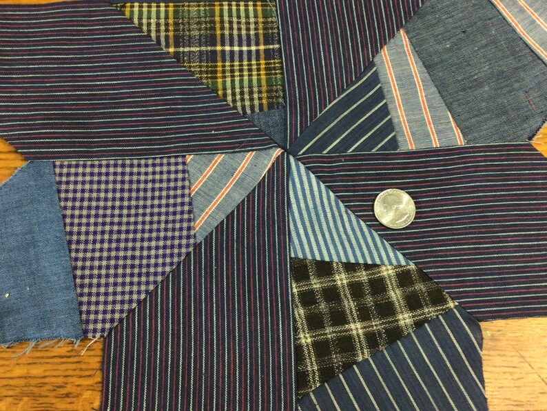 May include: A patchwork quilt block featuring a starburst design. The block is made from a variety of blue, white and black striped fabrics, as well as a plaid fabric and a gingham fabric. A silver coin is positioned in the centre of the block.