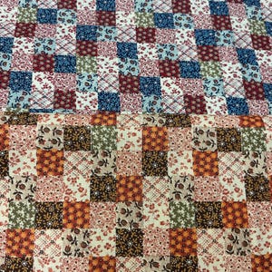Pode incluir: Um tecido com um design de patchwork de pequenos quadrados. A secção superior apresenta padrões florais em azul, vermelho e branco. A secção inferior mostra padrões florais em castanho, laranja e creme. O têxtil tem uma grelha repetitiva.