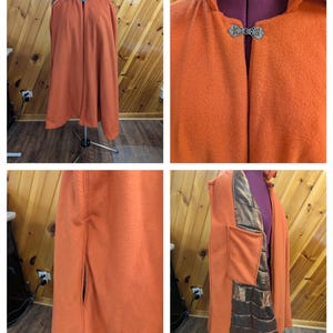 Può includere: Un mantello arancione con cappuccio e chiusura con fibbia decorativa. Il mantello è esposto su un manichino, mostrando la sua piena lunghezza e il suo design. Il mantello presenta una tasca e una fodera metallica.