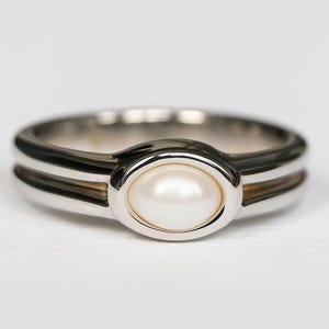 Könnte beinhalten: Silberring mit einem weißen, ovalen Perlmutt-Mittelstein. Der Ring hat ein Doppelband-Design mit poliertem Finish. Die Perle ist in einer Lünettenfassung gefasst, auf weißem Hintergrund.