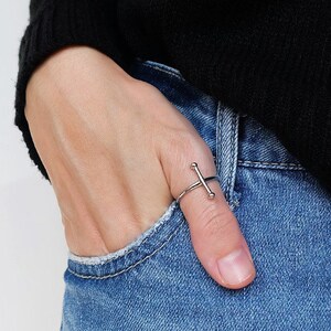May include: A silver ring with a simple cross design. The ring is worn on the thumb of a hand wearing blue jeans.