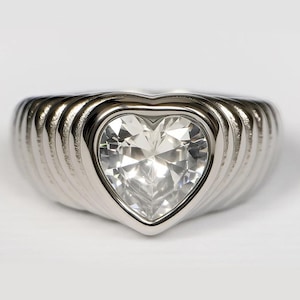 May include: A silver-toned ring with a heart-shaped, clear gemstone center. The ring has a ribbed, textured band. The gemstone is faceted, catching the light. The ring is set against a white background.