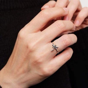 May include: A silver ring featuring a dragonfly design. The ring is worn on a person's finger.