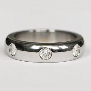 Op de afbeelding: Een gepolijste zilveren ring met drie heldere, ronde edelstenen gelijkmatig verdeeld over de band. De ring heeft een glad, afgerond ontwerp en reflecteert licht. De edelstenen zijn vlak met de band gezet.