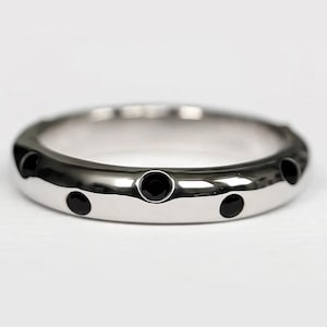 Puede incluir: Un anillo plateado pulido con una serie de piedras negras redondas incrustadas. El diseño es simple y elegante, con piedras espaciadas uniformemente alrededor del anillo.
